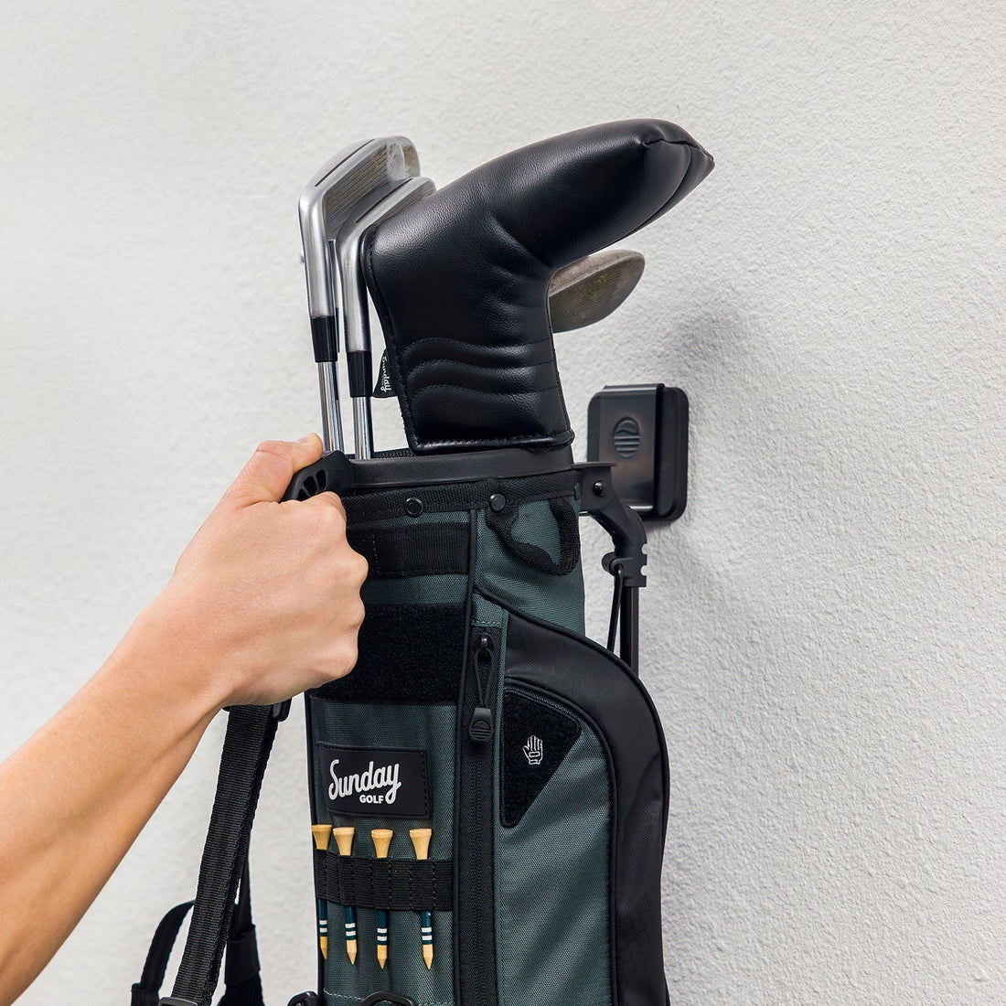 A hand placing a golf bag onto a black Sunday Golf Loma golf bag wall hook