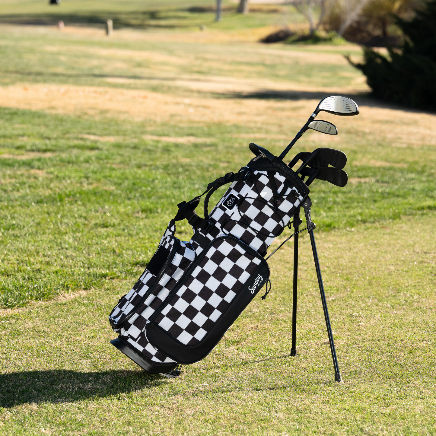 a checkered Black & White Recess kids golf bag with a stand, featuring multiple compartments and golf clubs inside, set on a golf course