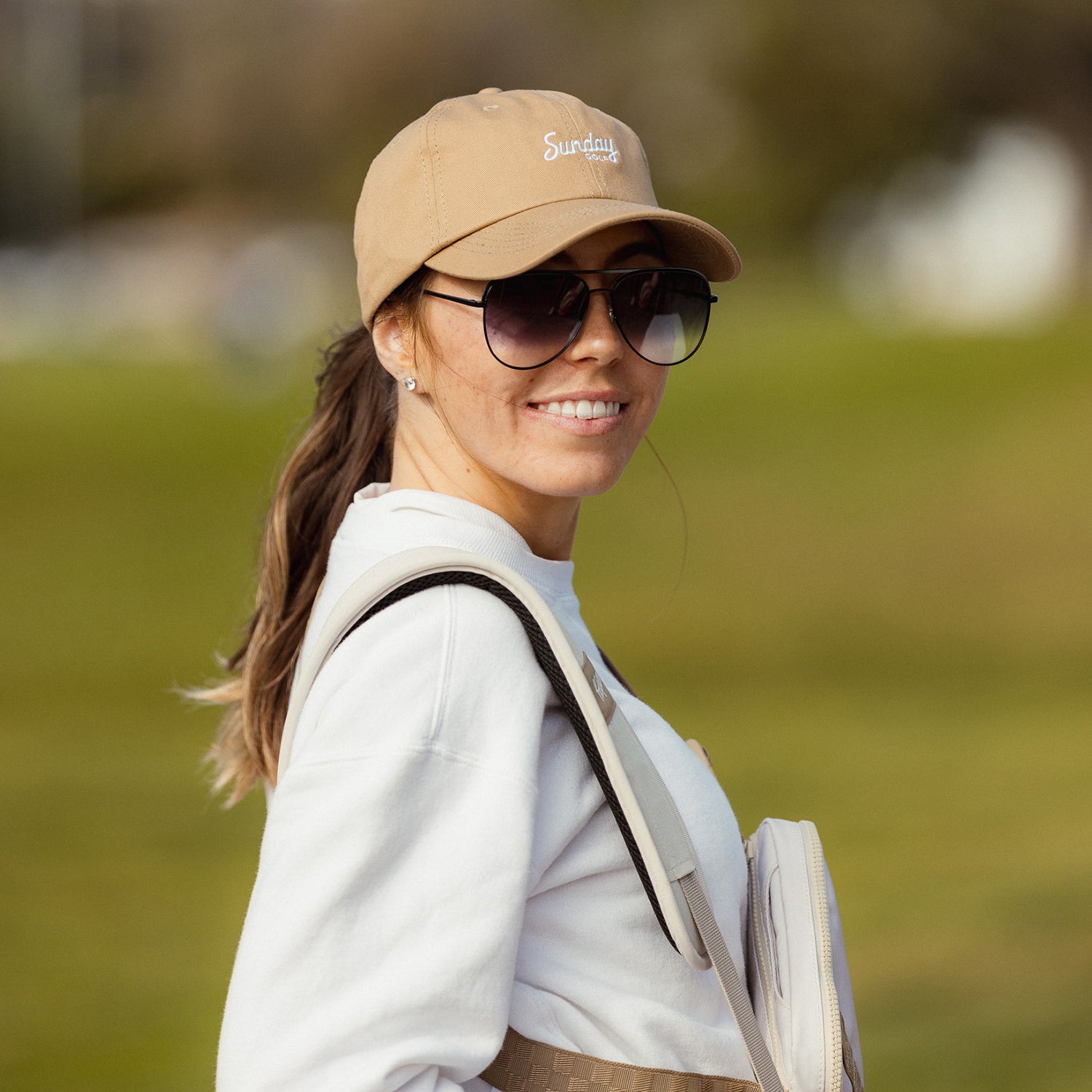 A woman smiling at the camera while wearing khaki dad golf hat and sunglasses