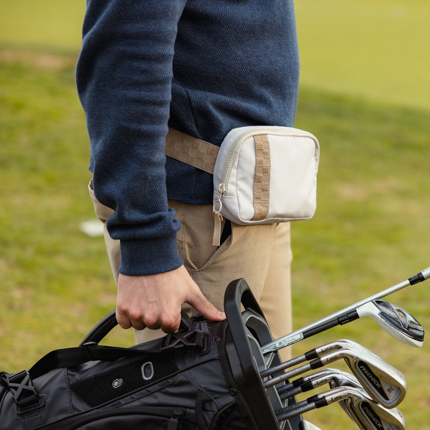 A man wearing a toasted almond belt bag on his waist while holding a golf bag with golf clubs in one hand