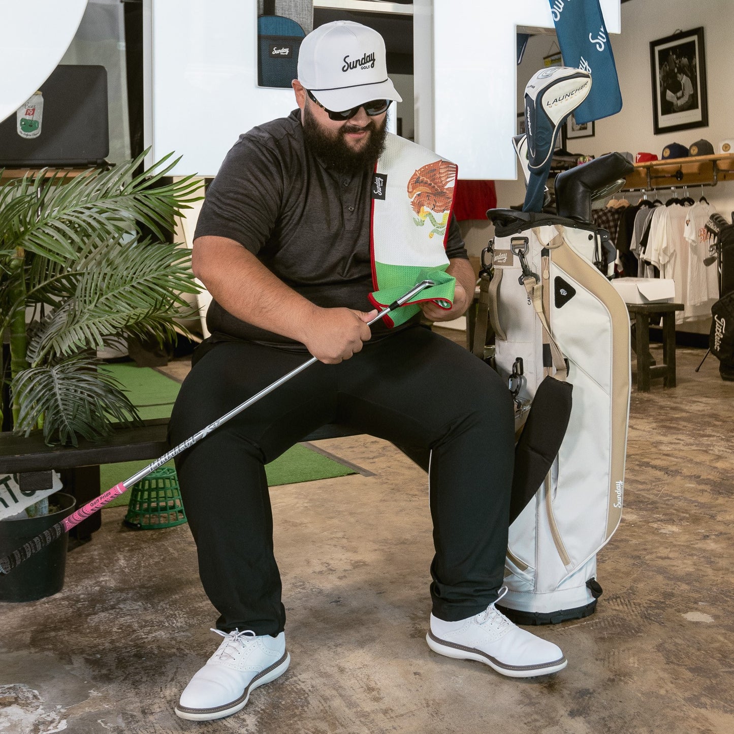 A man wearing white and black rope golf hat while cleaning his clubs with golf towel and sitting beside his toasted almond golf bag