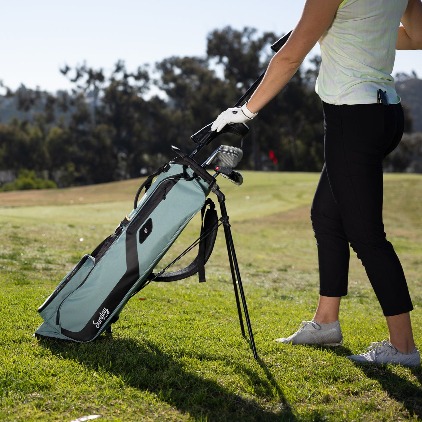 A woman putting clubs in the seafoam green El Camino golf bag standing on the course