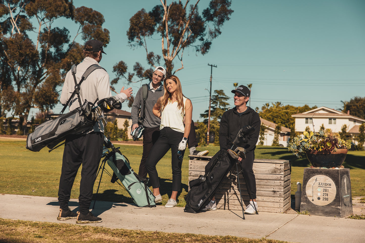 A man standing with heather gray El Camino golf bag, a woman standing with seafoam El Camino golf bag, and a man sitting with matte black El Camino golf bag