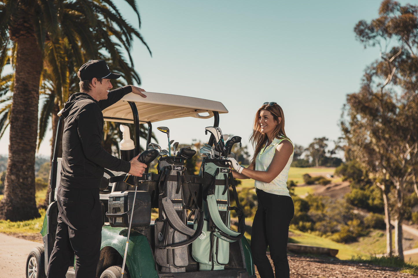 A man with heather gray El Camino golf bag talking to a woman with seafoam El Camino golf bag on a golf cart