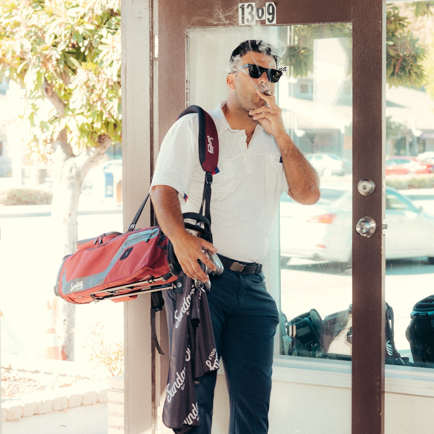 A man smoking a cigar carrying El Camino golf bag in deep red burgundy with a hanging Sunday Golf Towel