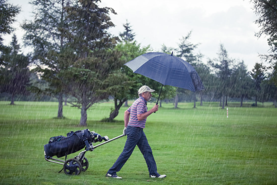 A golfer walking on the course while raining while holding an umbrella and strolling his golf bag