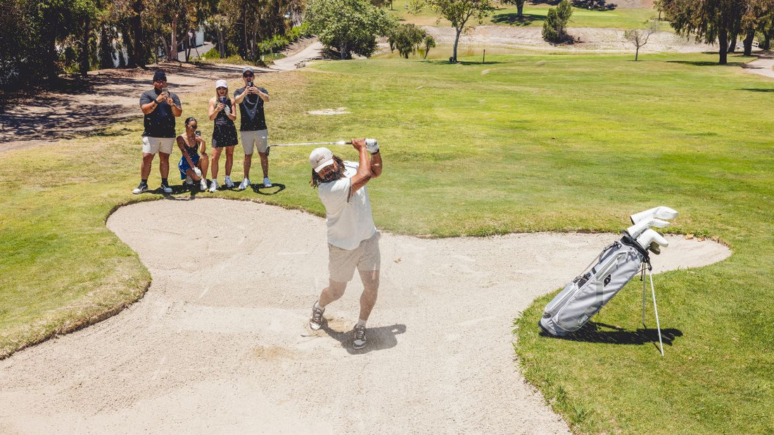 A golfer swinging out of a sand bunker while friends watch and film