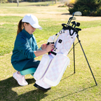A young girl wearing a white cap and teal jacket kneels on a golf course, adjusting the straps of a checkered Pink & White Recess kids golf bag with a stand. The bag is equipped with multiple compartments and golf clubs inside