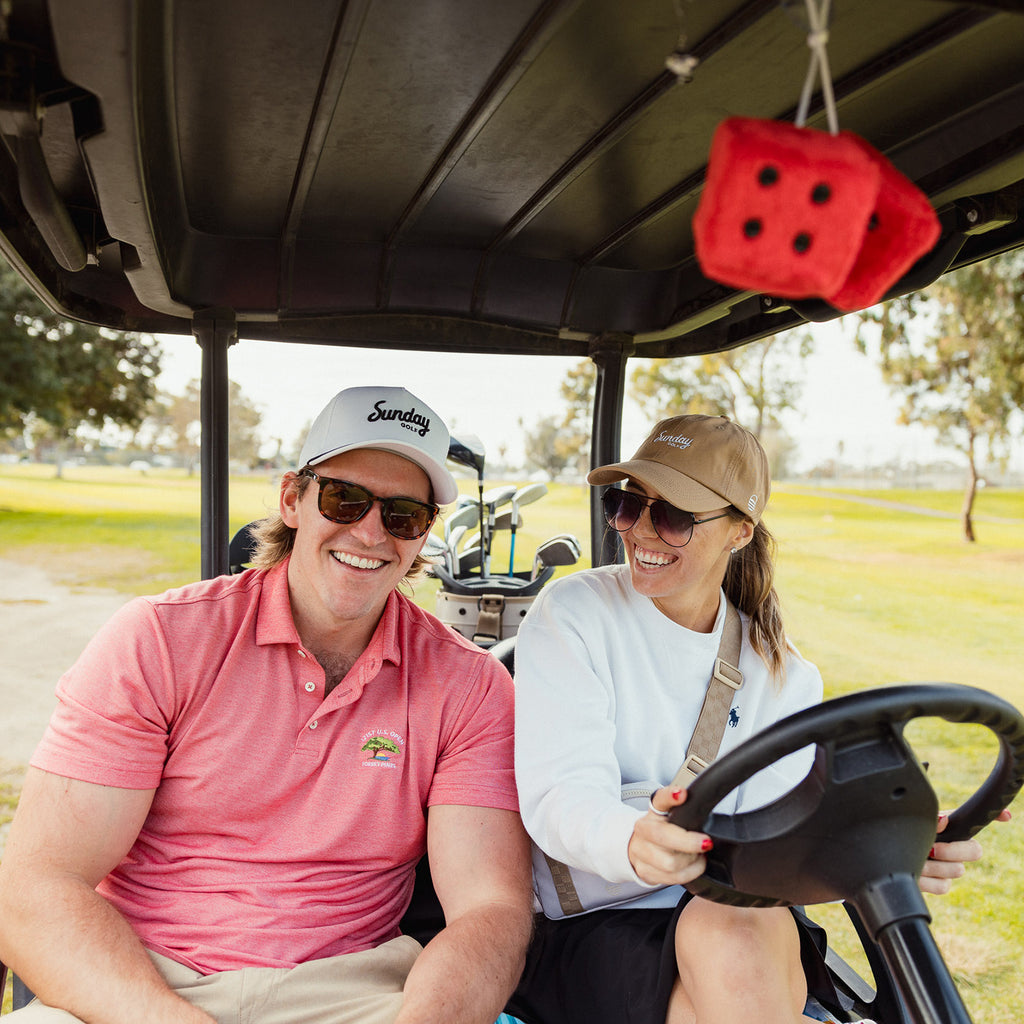 A man wearing white and black rope golf hat on the passenger seat beside a woman wearing a dad golf hat while driving a golf cart