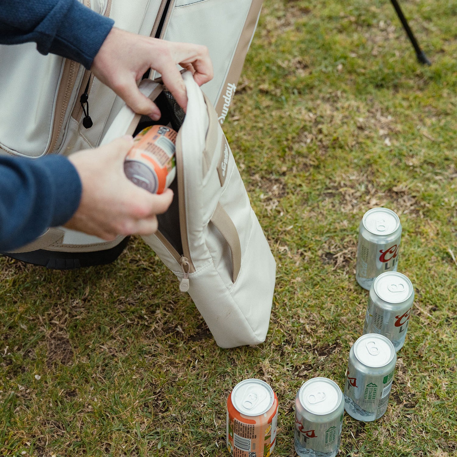 A canned drink being placed inside a toasted almond big frosty golf cooler bag on the ground with a couple more cans beside it
