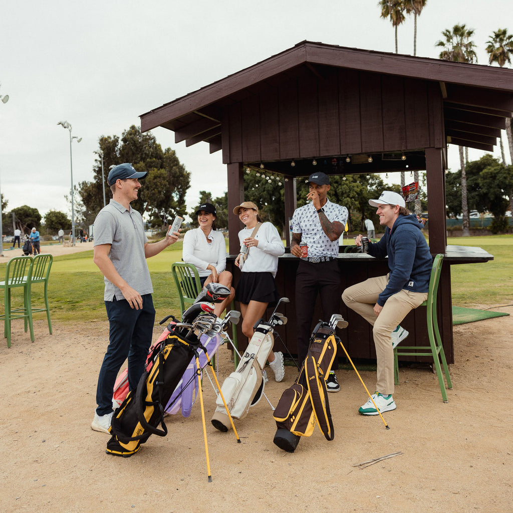 A group of people laughing and holding canned drinks beside their golf bags and one of them is an El Camino in black and yellow