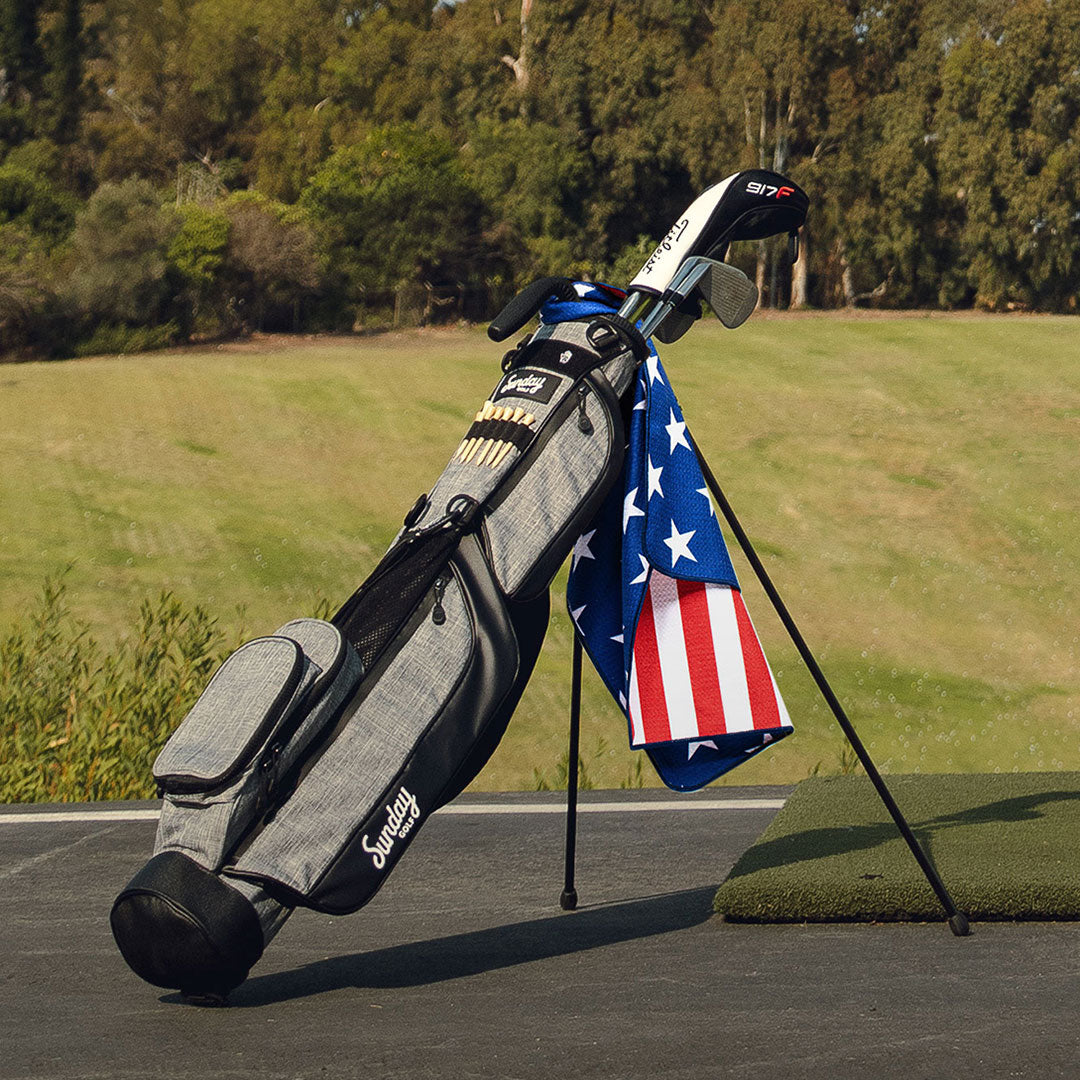 A full front shot of standing Loma golf bag in heather gray with an American flag golf towel hanging on it