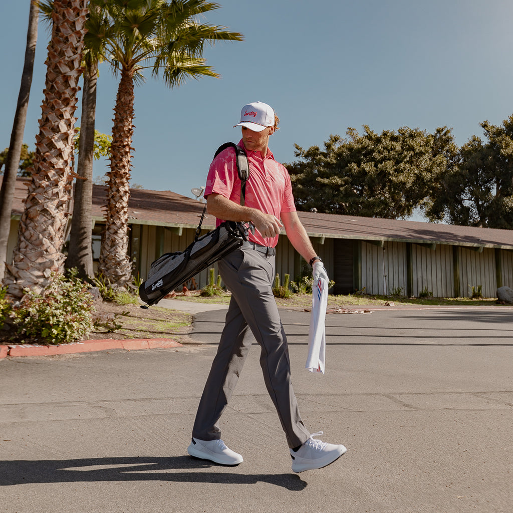 A man wearing tropical rope golf hat carrying gray Loma golf bag and el classico golf towel by Sunday Golf