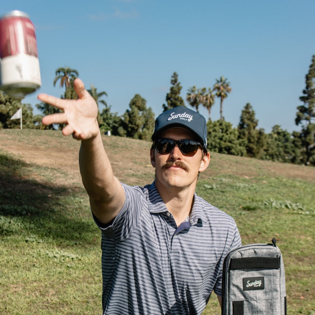 A man tossing beverage wearing navy rope golf hat