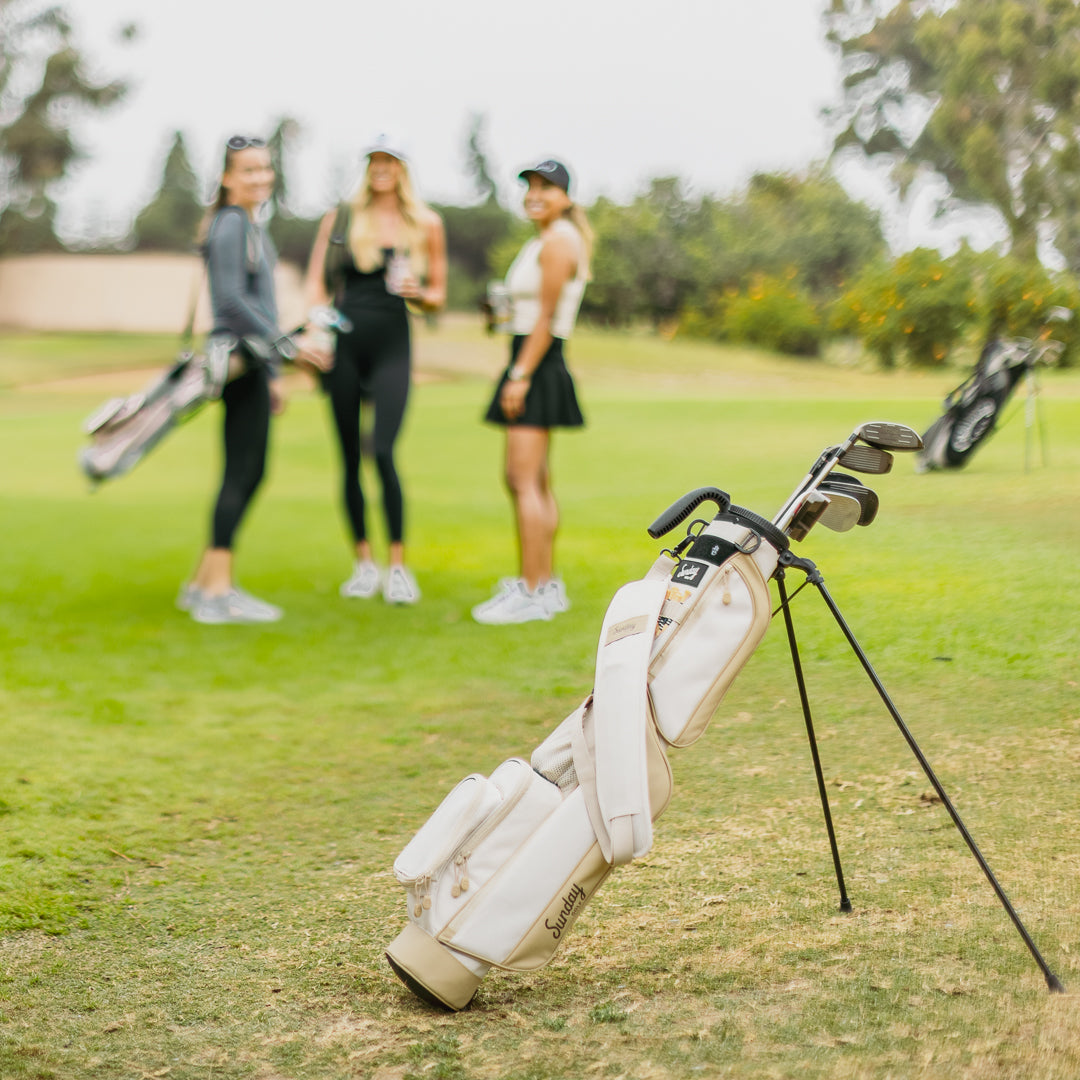 A full front shot of standing Loma golf bag in toasted almond with single strap on the course with three women standing in the background