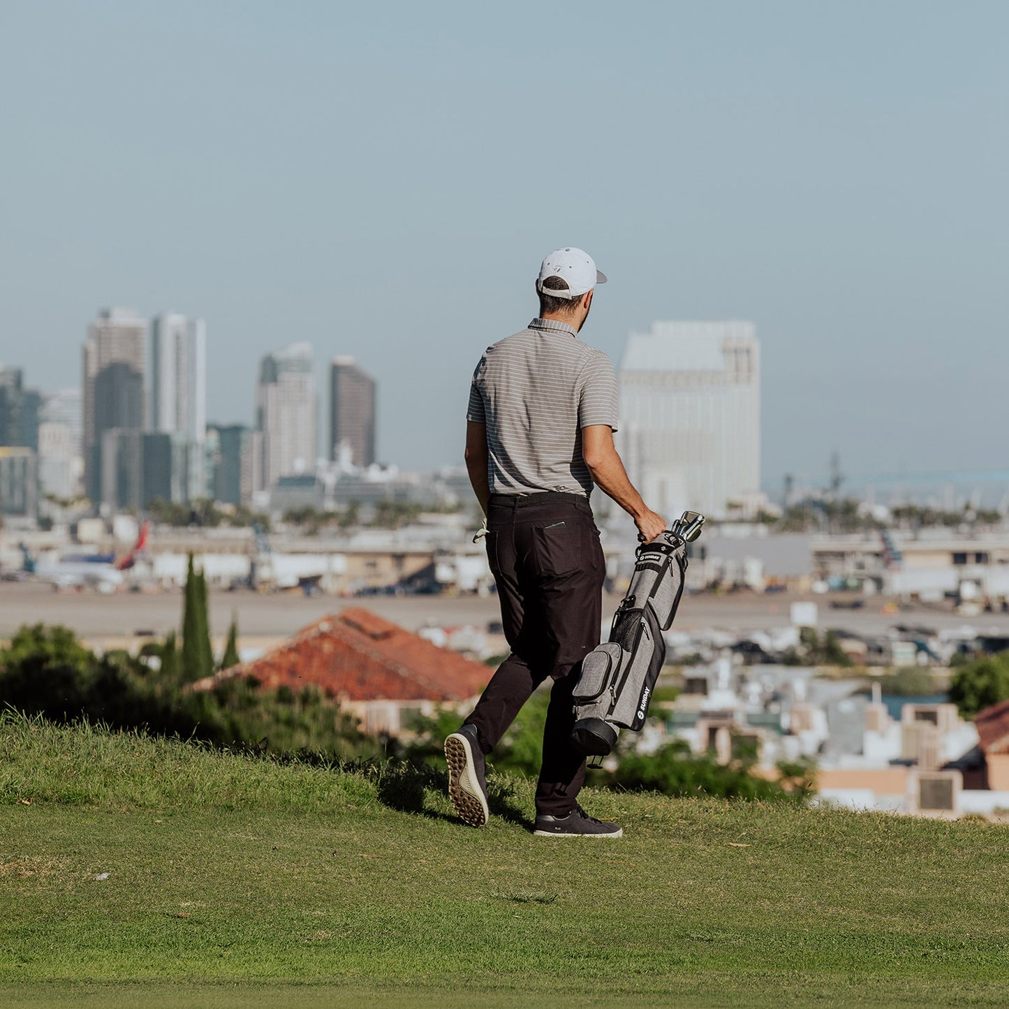 A golfer carrying a heather gray Loma golf bag with one hand