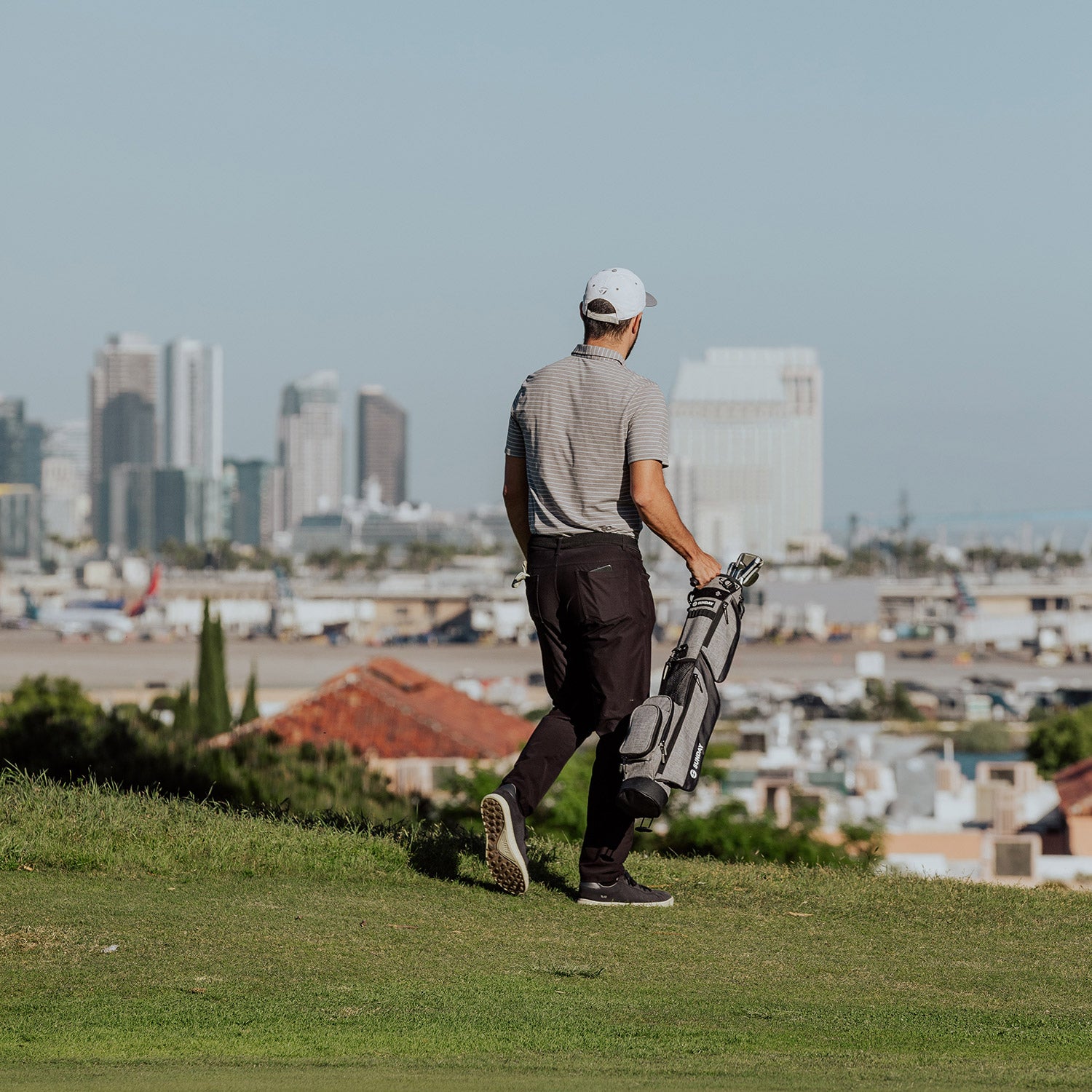 A golfer carrying a heather gray Loma golf bag with one hand