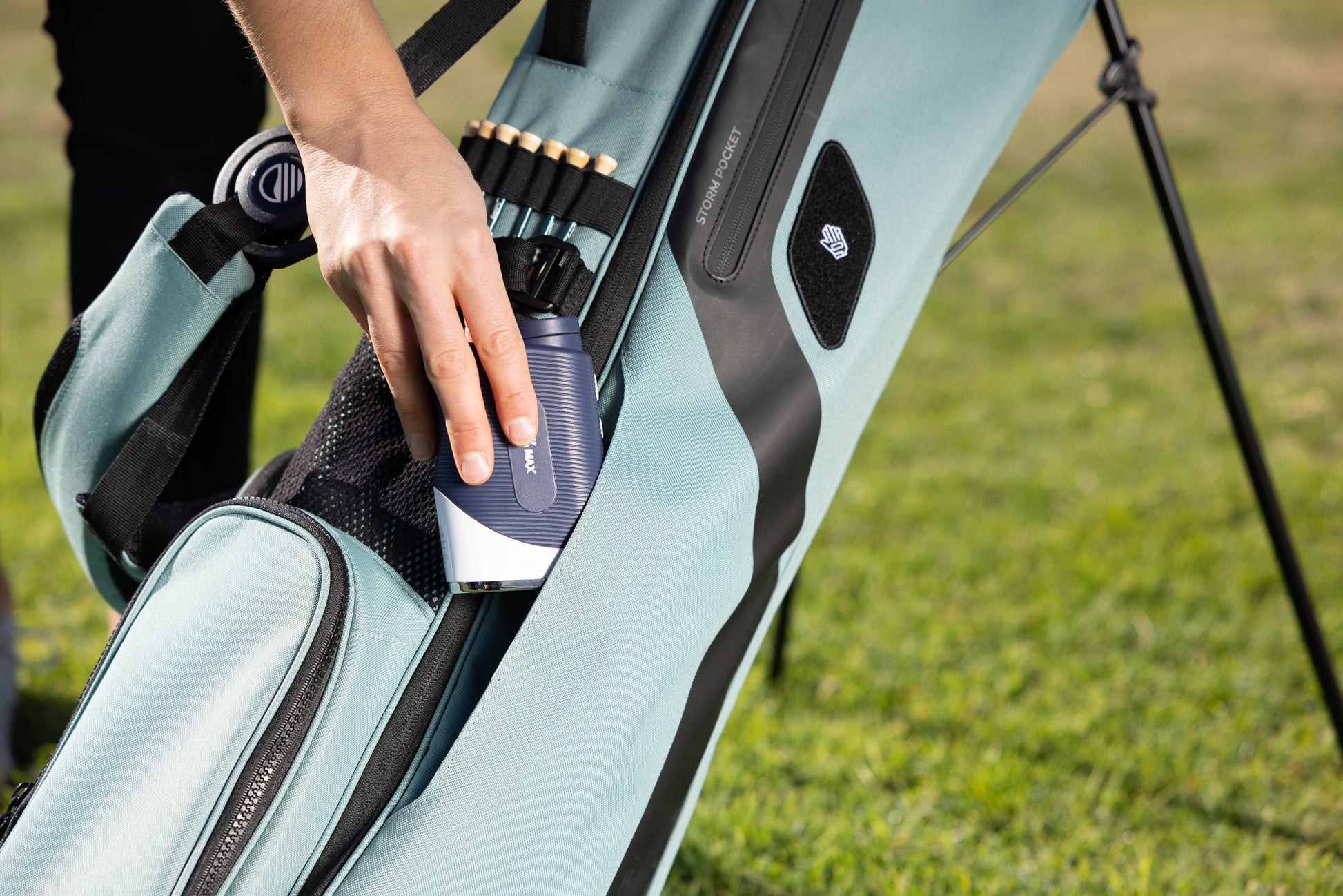 A close up shot of a woman putting a range finder into seafoam green El Camino golf bag's side pocket