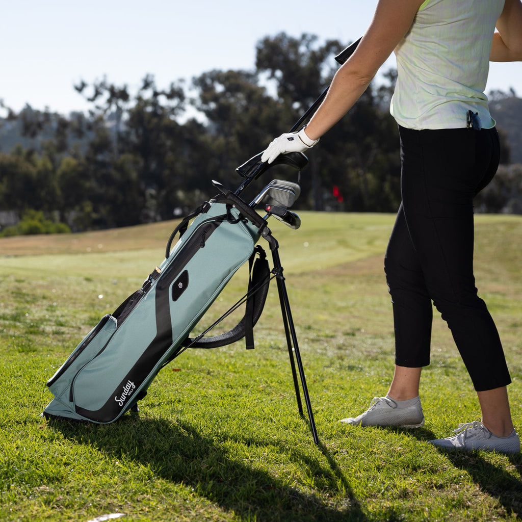 A woman putting clubs in the seafoam green El Camino golf bag standing on the course