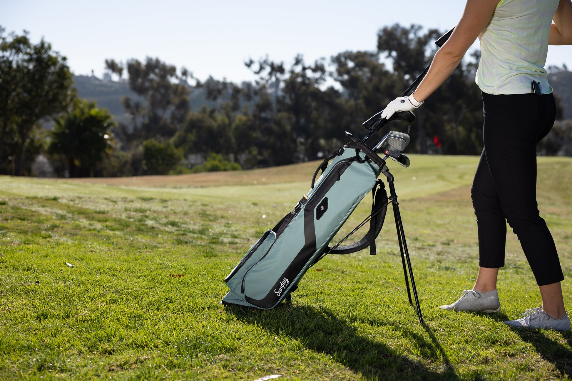 A woman putting clubs in the seafoam green El Camino golf bag standing on the course