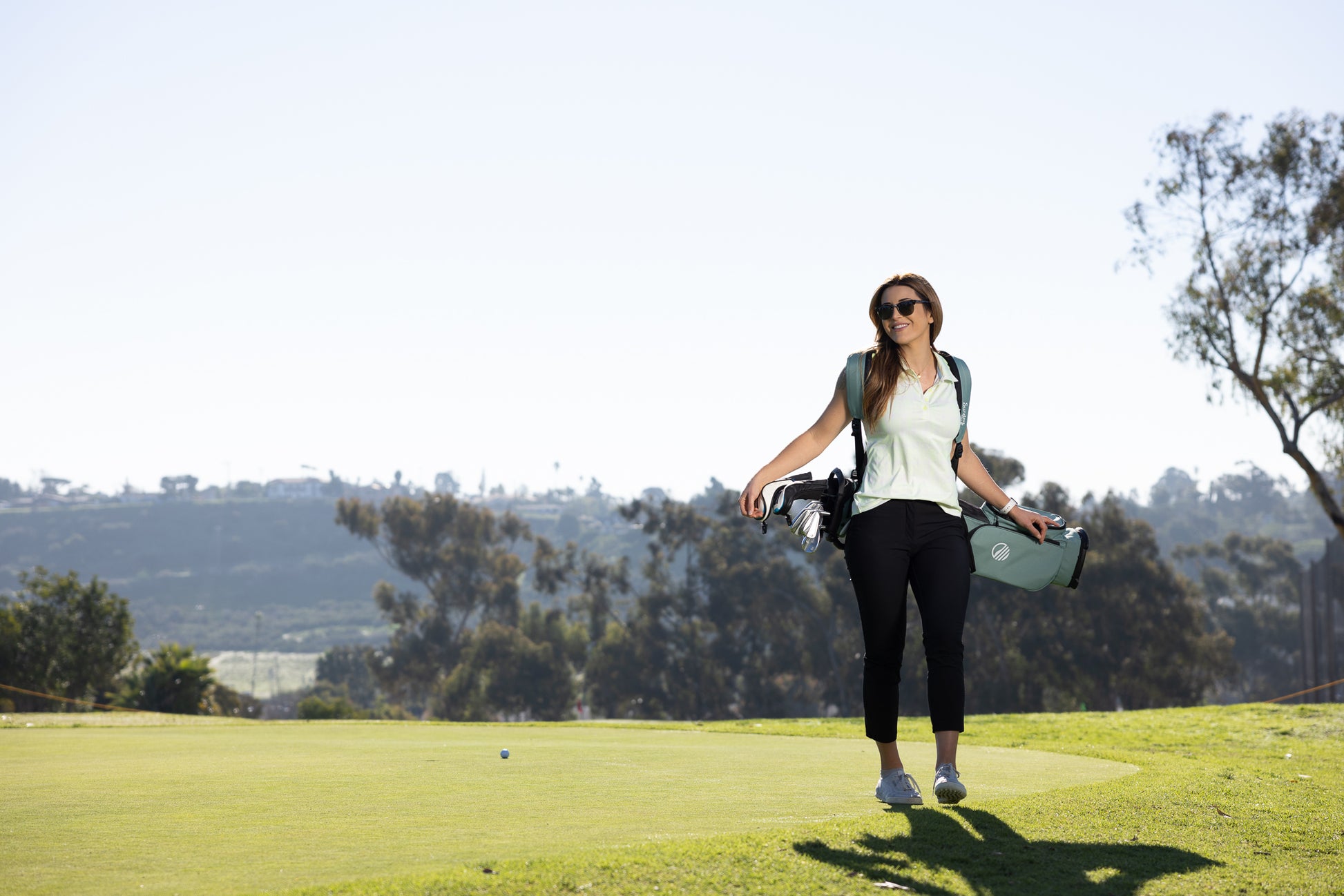 A woman smiling, carrying seafoam green El Camino golf bag while standing on the course