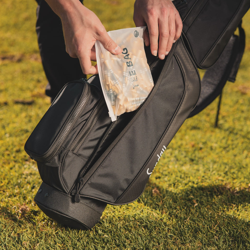 A tee bag of bamboo golf tees being placed inside a black golf bag