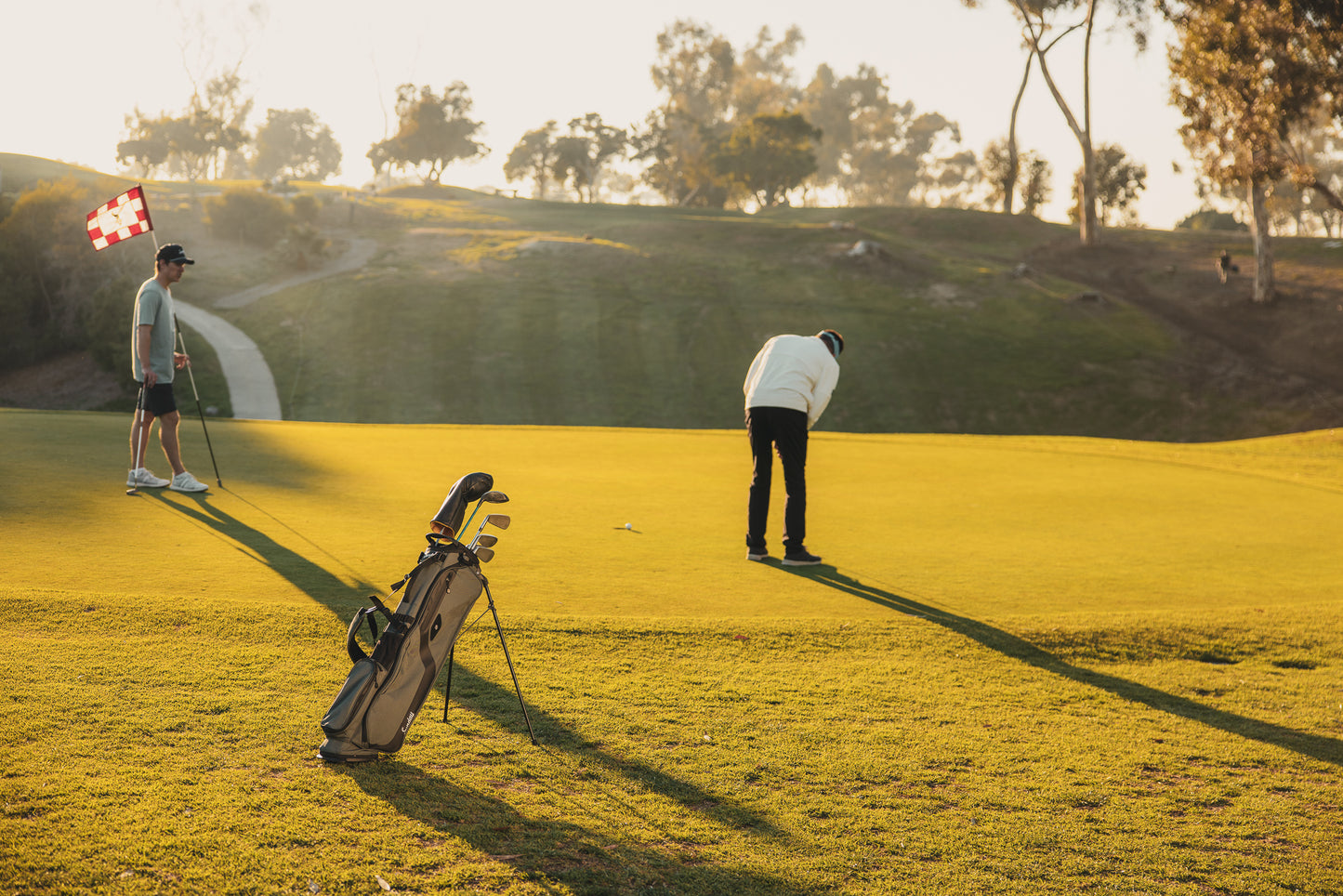 A standing El Camino Golf bag in midnight green on golf course with two golfers on the green putting 