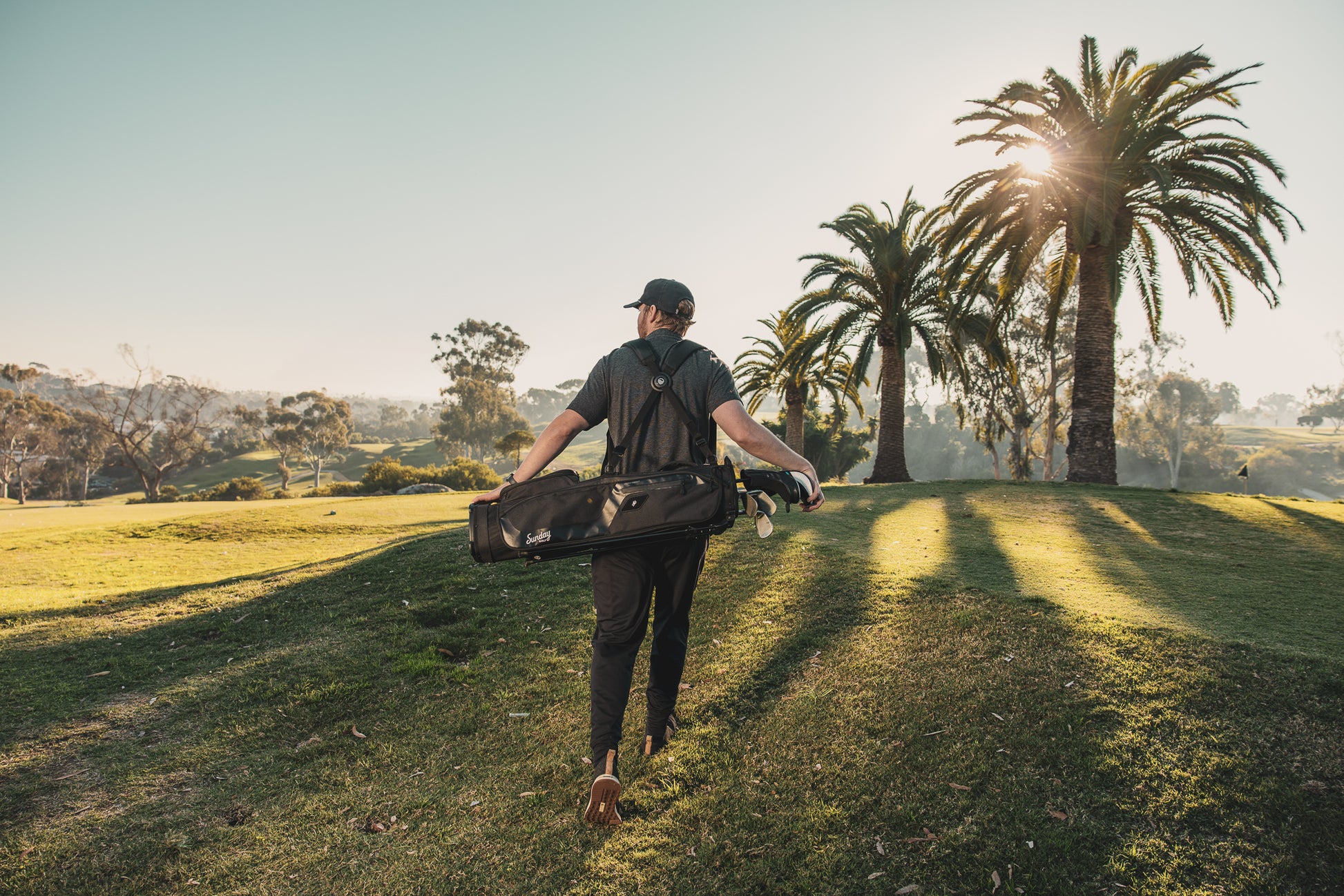 man carrying the matte black El Camino golf bag by Sunday golf on golf course with palm trees