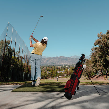 A man swinging a golf club at a driving range with a deep red burgundy Loma XL golf bag standing behind him
